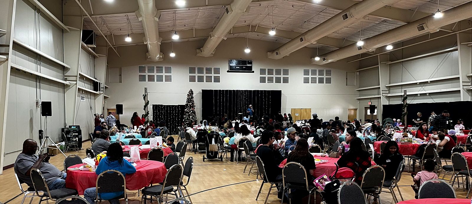 Families at tables with red tablecloths in the gym, a Christmas tree in the corner
